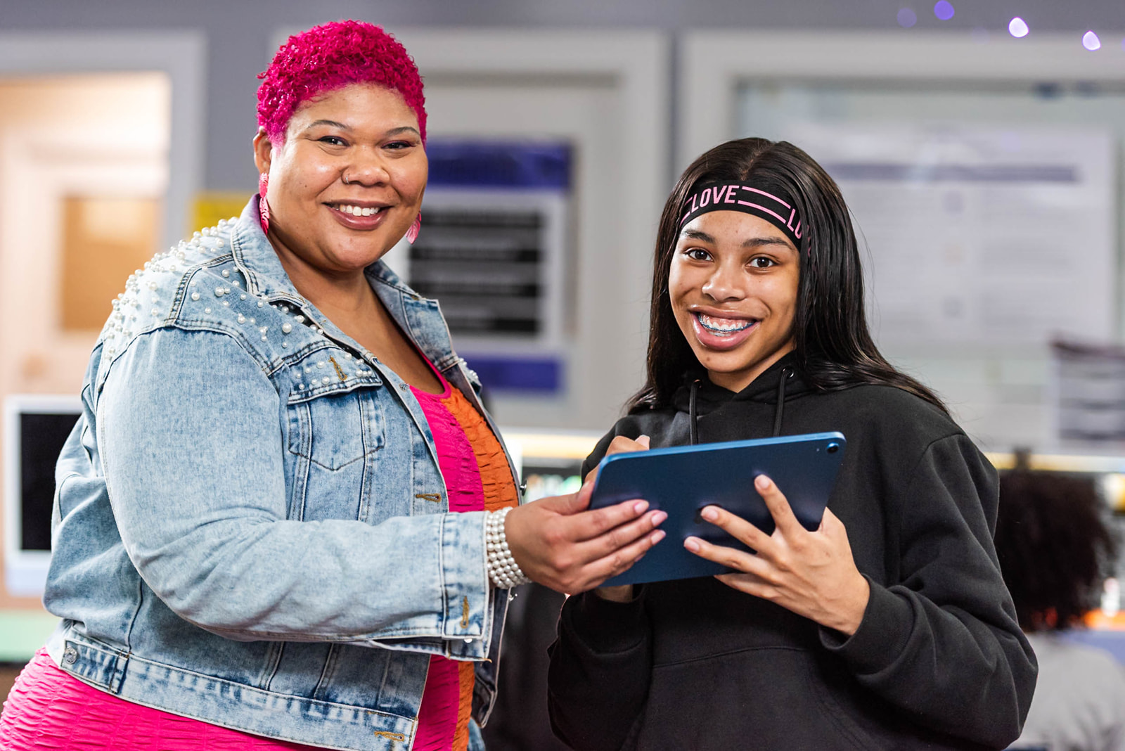 An older woman with bright pink hair and a younger girl with braces stand together in a classroom, smiling while holding and looking at a tablet.