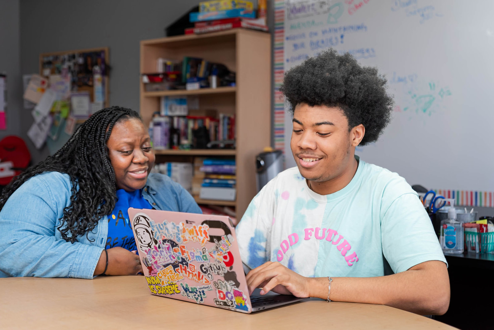 A young man and an older woman sit at a table in a classroom, smiling as they look at a sticker-covered laptop together, with bookshelves and a whiteboard in the background.