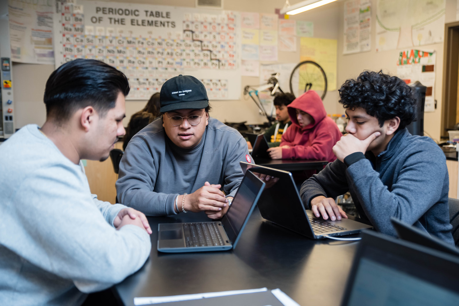 Three students sit around a table in a classroom focused on their laptops with a periodic table visible on the wall behind them and another two students working in the background.