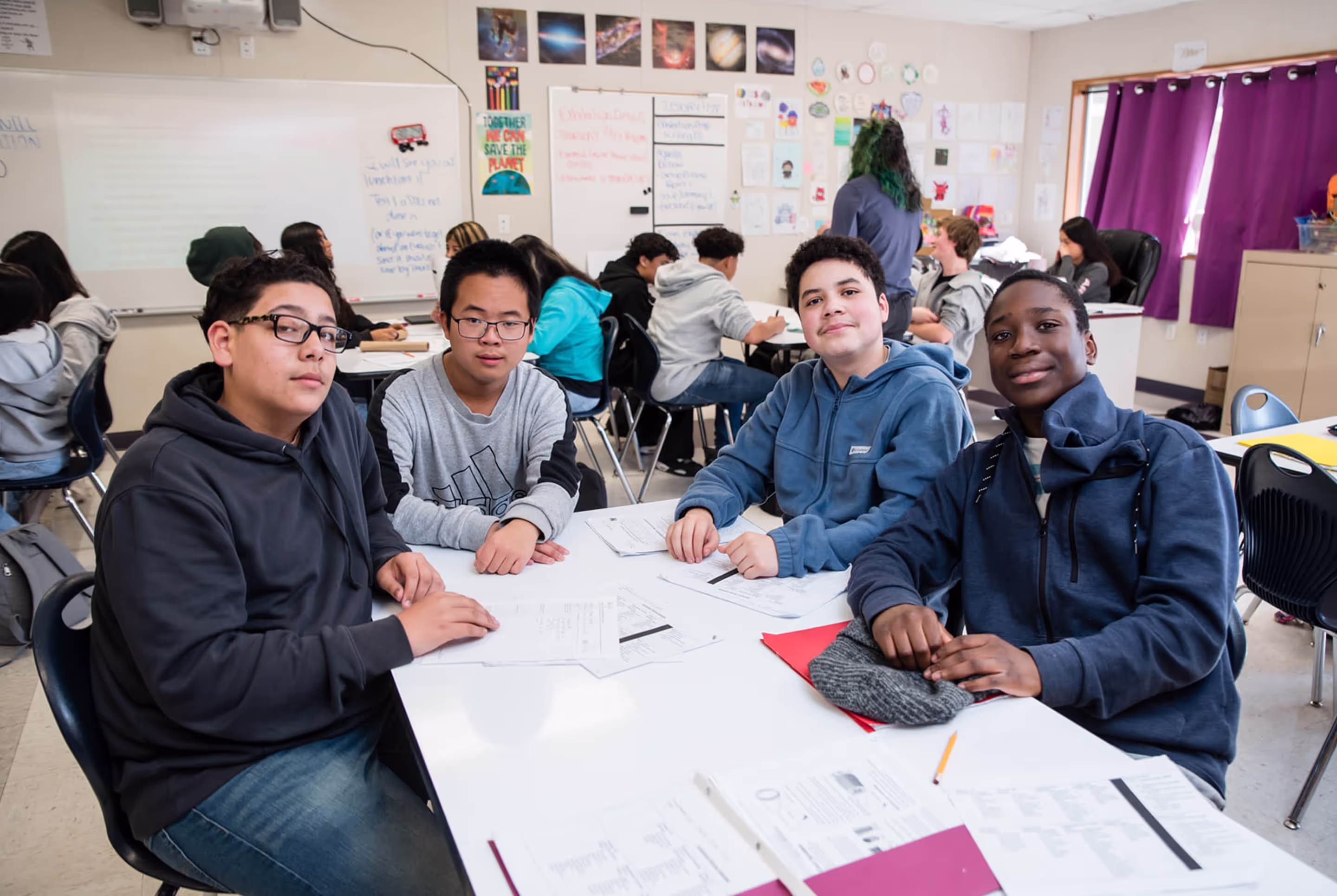 Four teenage boys seated at a classroom table with papers, looking at the camera; other students work in groups in the background with colorful posters on the walls.