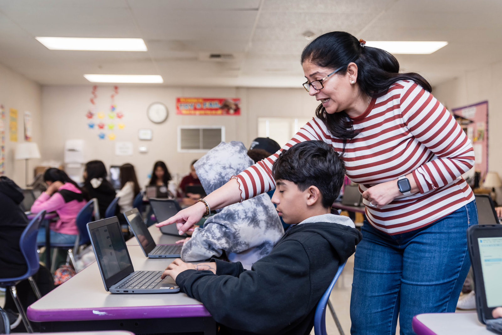 Teacher in a striped shirt helping a student working on a laptop in a classroom with other students using laptops.