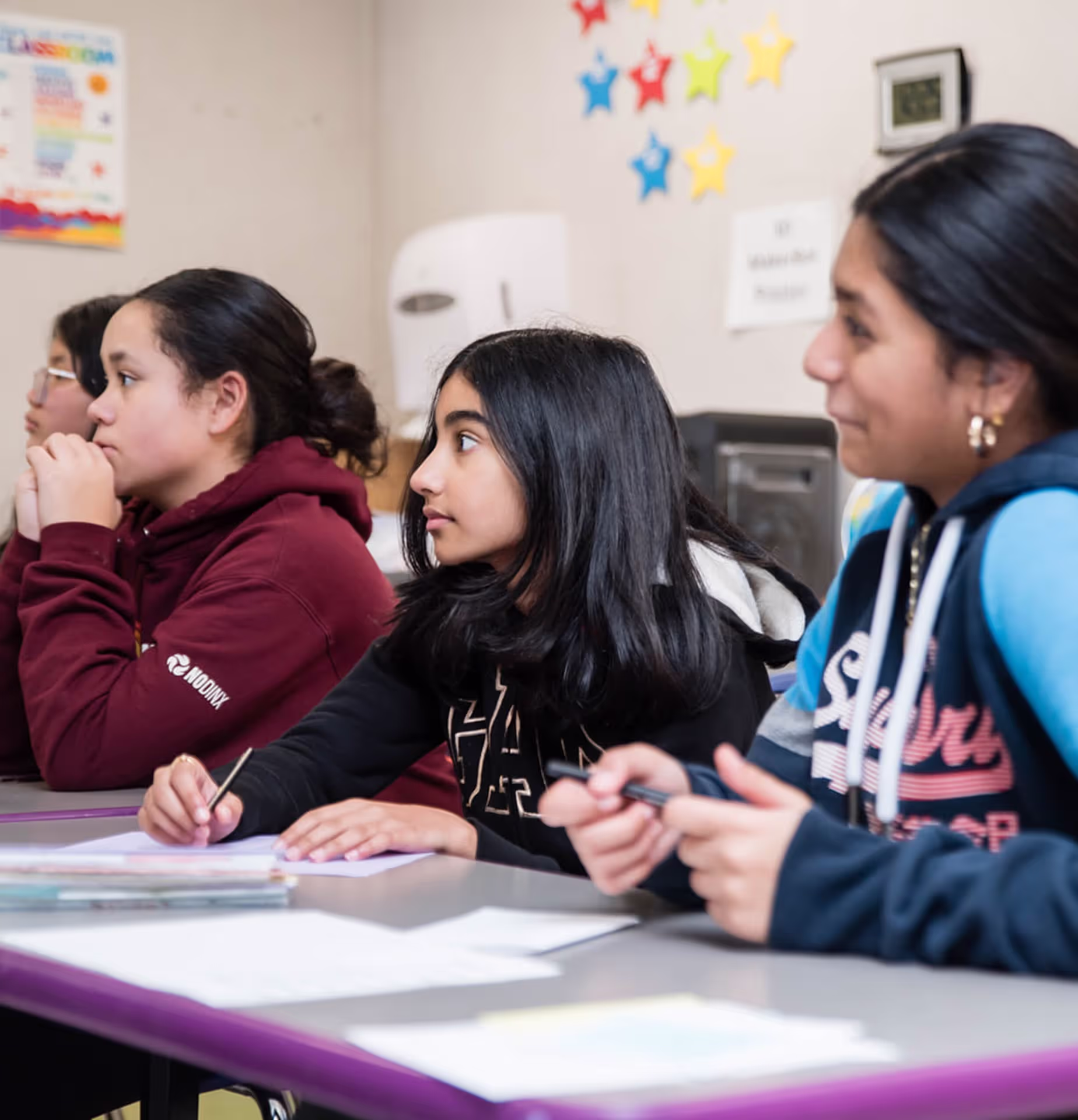 Four students seated at desks in a classroom, attentively listening and holding pens with papers in front of them.