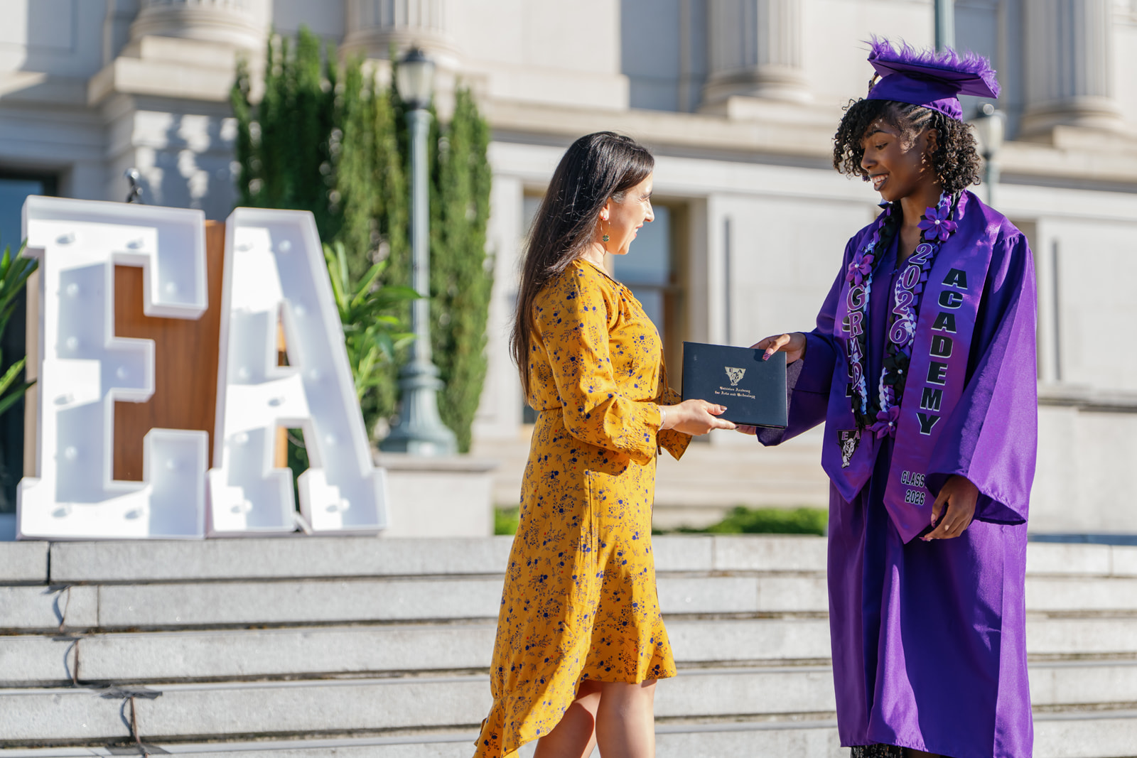 Woman in a purple graduation gown and cap receiving a diploma from another woman in a yellow floral dress outdoors on steps with large lit letters 'EA' in the background.