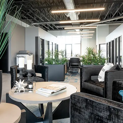 Modern office lobby with black velvet armchairs, round coffee tables, decorative books, candles, and indoor plants under linear ceiling lights.