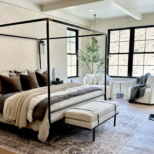 Modern bedroom with a black metal canopy bed, layered neutral bedding, a cushioned bench, two white armchairs, large windows, and a potted tree.