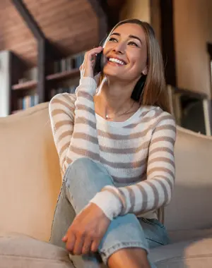 Smiling woman sitting on a couch talking on a smartphone in a cozy living room.