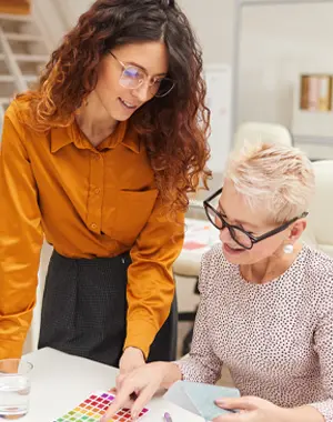 Two women, one standing and one seated, reviewing color swatches and fabric samples in a bright workspace.