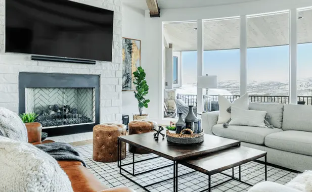 Bright living room with large windows showing mountain view, gray sofa, leather chair, coffee tables, and a TV above a white brick fireplace.