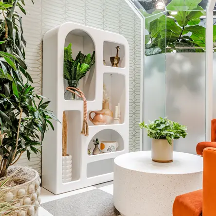 Modern interior featuring a white uniquely shaped shelf with plants and decor, a round white table with a potted plant, and orange chairs.