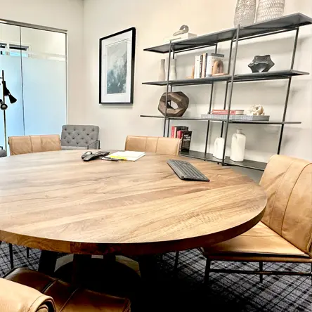 Round wooden meeting table with a keyboard, phone, and papers surrounded by four brown leather chairs in a modern office with a shelving unit and framed artwork.