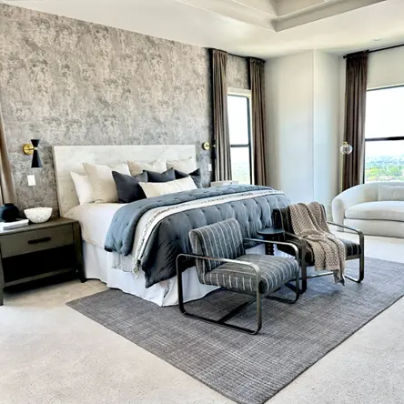 Modern bedroom with a large bed, gray textured accent wall, two striped armchairs on a gray rug, and large windows with brown curtains.