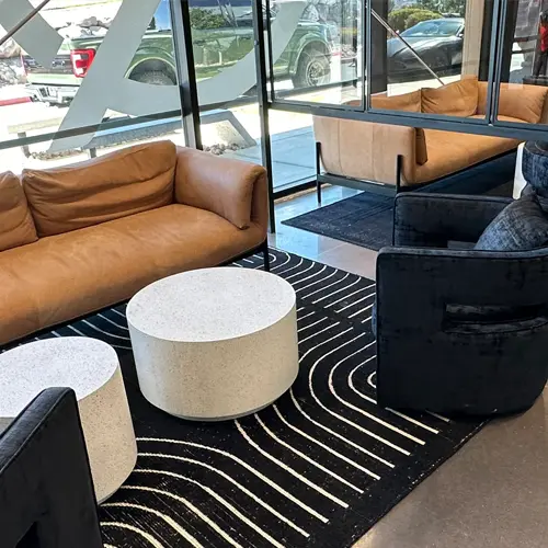 Modern seating area with a brown leather sofa, black armchairs, two round white coffee tables, and a black rug with white wavy lines near large glass windows.