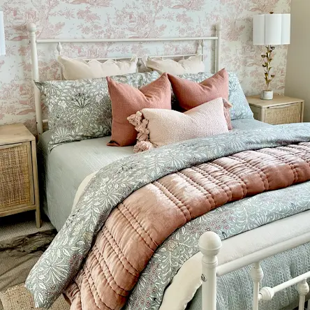 Cozy bedroom with white metal bed frame, floral patterned bedding in soft gray and blush pink pillows, beside wicker nightstands with a lamp.