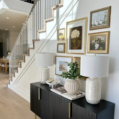Modern staircase with white railing beside a black console table displaying two white textured lamps, a decorative vase with green leaves, and framed floral artwork on the wall.