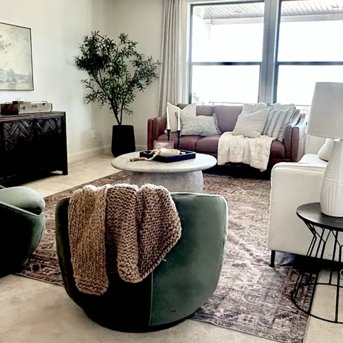 Cozy living room with a brown sofa, white armchair, green velvet chair with a knitted blanket, round coffee table, and a large window providing natural light.