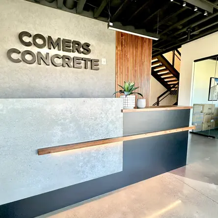 Modern office reception area with a concrete and wood desk, a plant on the counter, and wall signage reading 'COMERS CONCRETE'.