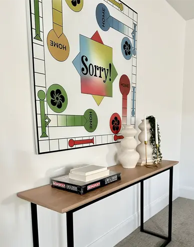Modern console table with a Sorry board game mounted on the wall above, decorated with two white vases and wine glasses on the table.