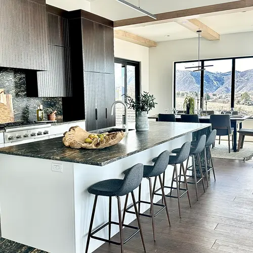 Modern kitchen with dark wood cabinets, a large island with dark stone countertop, six gray bar stools, and large windows showing a mountainous view.