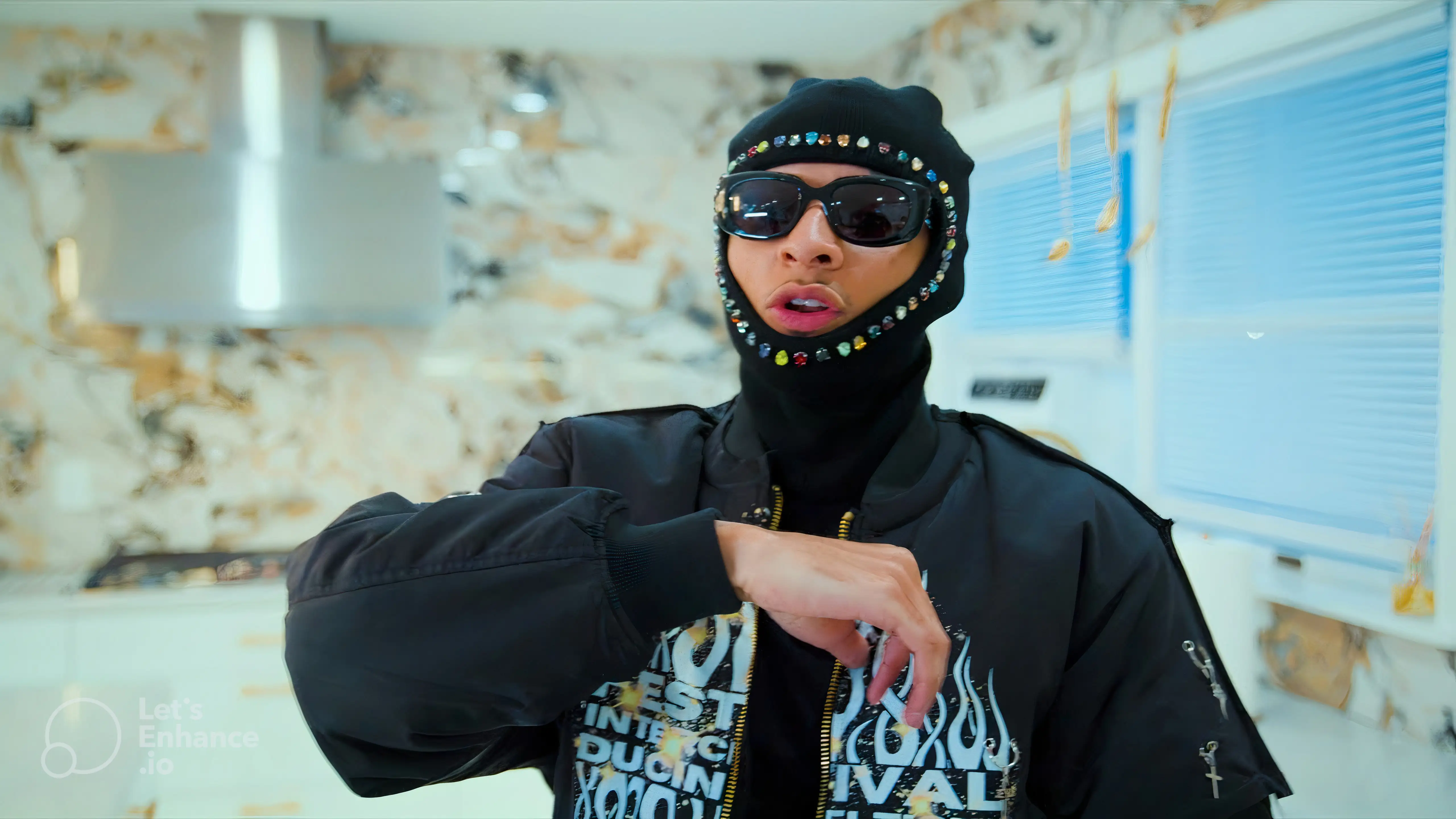 Person wearing a black jacket with white text and designs, a black balaclava adorned with colorful gems, and large sunglasses inside a room with blue blinds.
