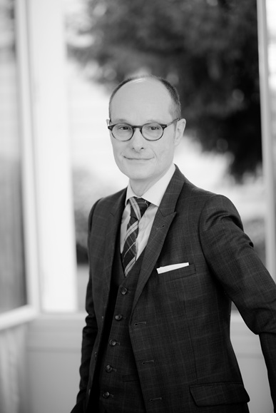 Homme portant une veste noire et une chemise blanche, appuyé sur un meuble avec un fond de rideaux clairs.