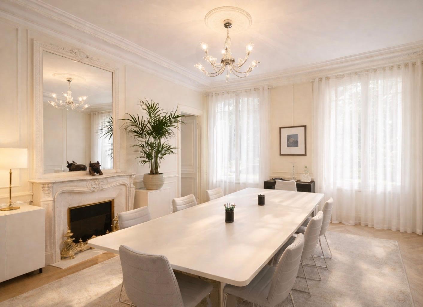 Salle de réunion lumineuse avec une grande table blanche, huit chaises grises, un grand miroir, une cheminée décorative et un parquet en chevrons.
