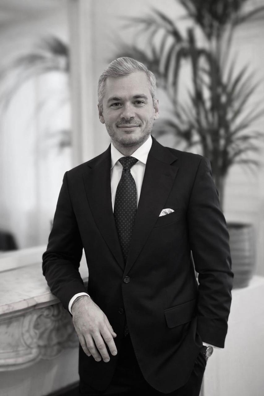 Homme en costume sombre et cravate assis, regardant vers l'avant avec une légère expression souriante.