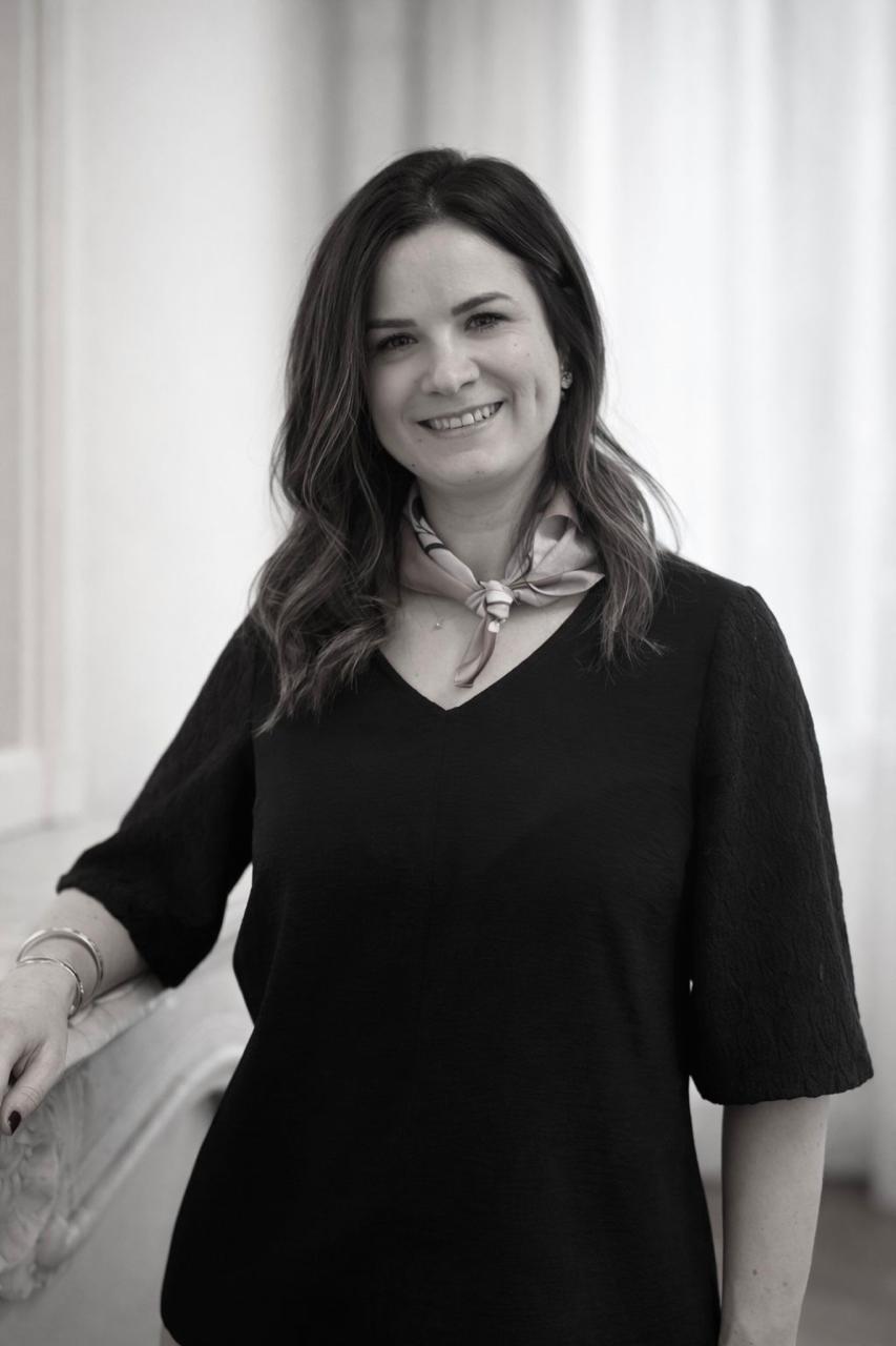 Portrait en noir et blanc d'une femme aux cheveux longs et droits, souriante, bras croisés, portant un haut à manches longues.