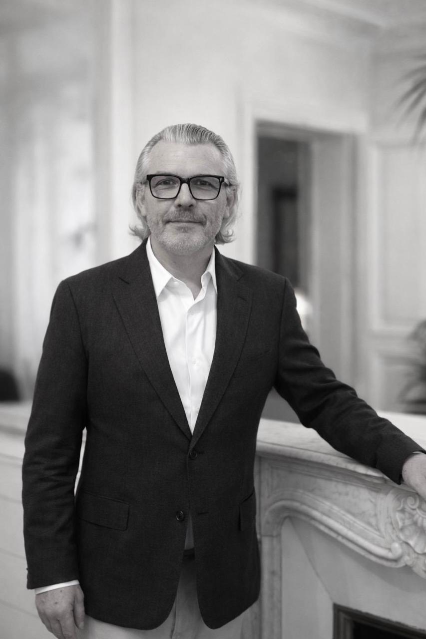 Homme portant une veste noire et une chemise blanche, appuyé sur un meuble avec un fond de rideaux clairs.