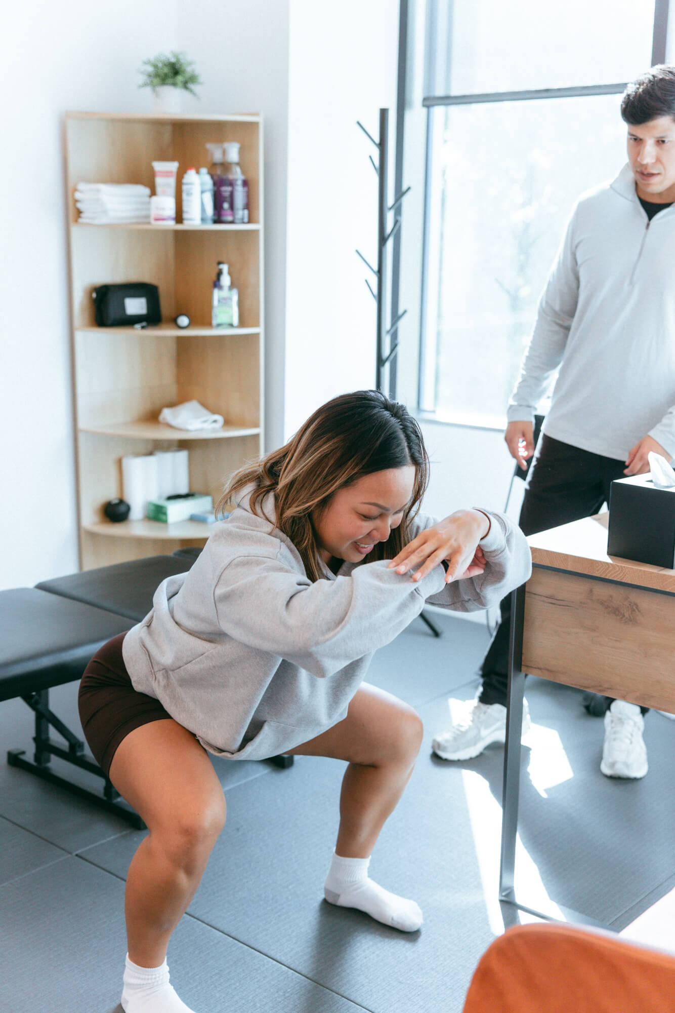 Woman in gray hoodie performing a squat exercise indoors while a man in light gray stands nearby watching.