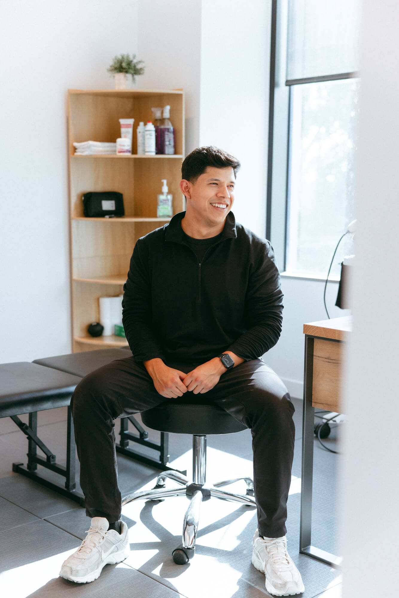 Smiling man in black pullover and white sneakers sitting on a swivel stool in a bright room with wooden shelf and window.