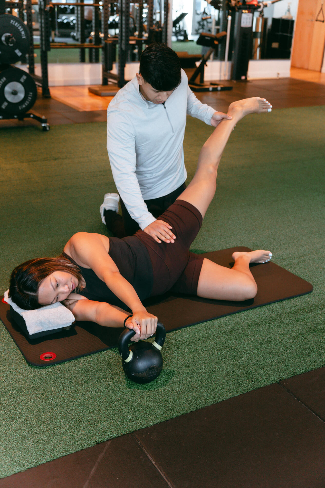 A female client on a black mat holds a kettlebell while a male trainer supports her raised leg during a gym stretching exercise.