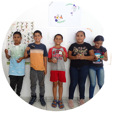 Five children standing against a white wall with colorful drawings, holding small craft items.