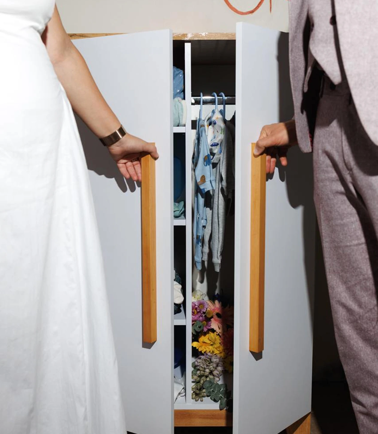 Два человека держат деревянные ручки шкафа, внутри котором висят детские вещи и лежат цветы.