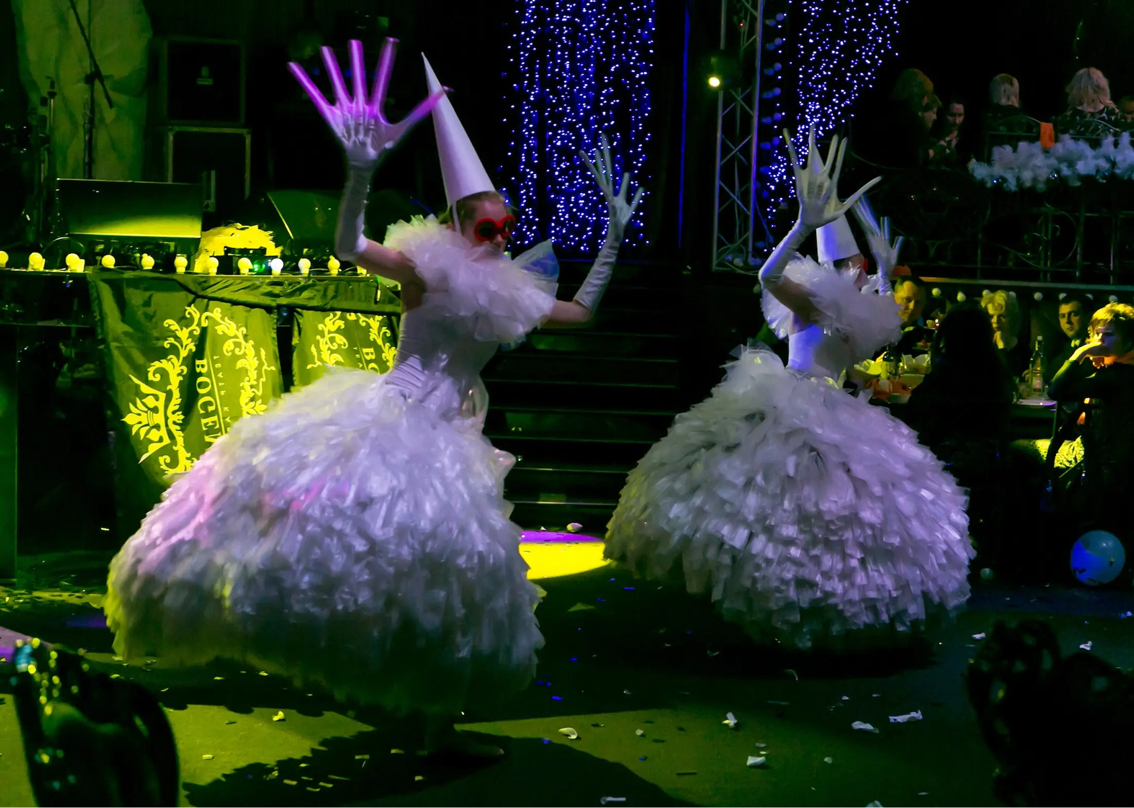 Two performers in white ruffled dresses, white cone hats, long gloves, and red glasses dancing at a colorful stage event.