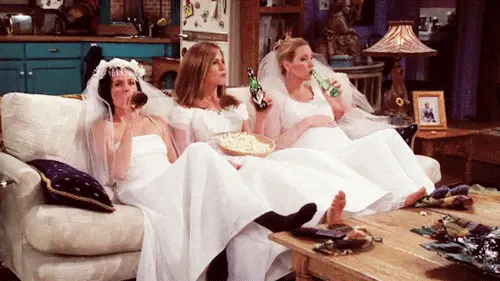 Three women in wedding dresses sitting on a couch drinking beer and eating popcorn in a cozy living room.