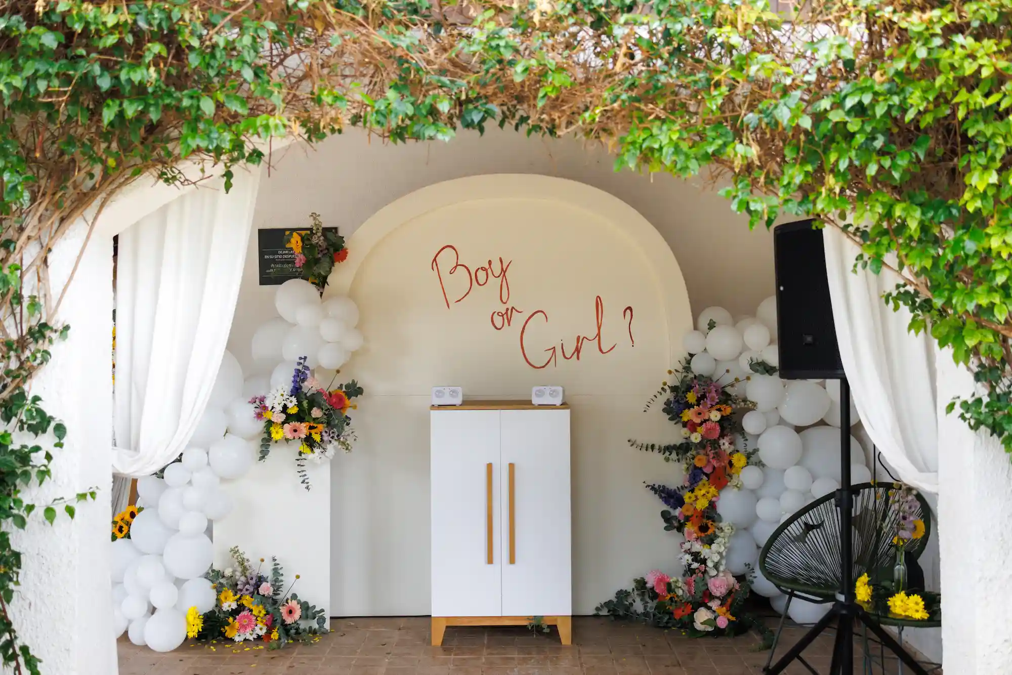 Decorated archway with white balloons, colorful flowers, and a sign reading 'Boy or Girl?' for a gender reveal party.