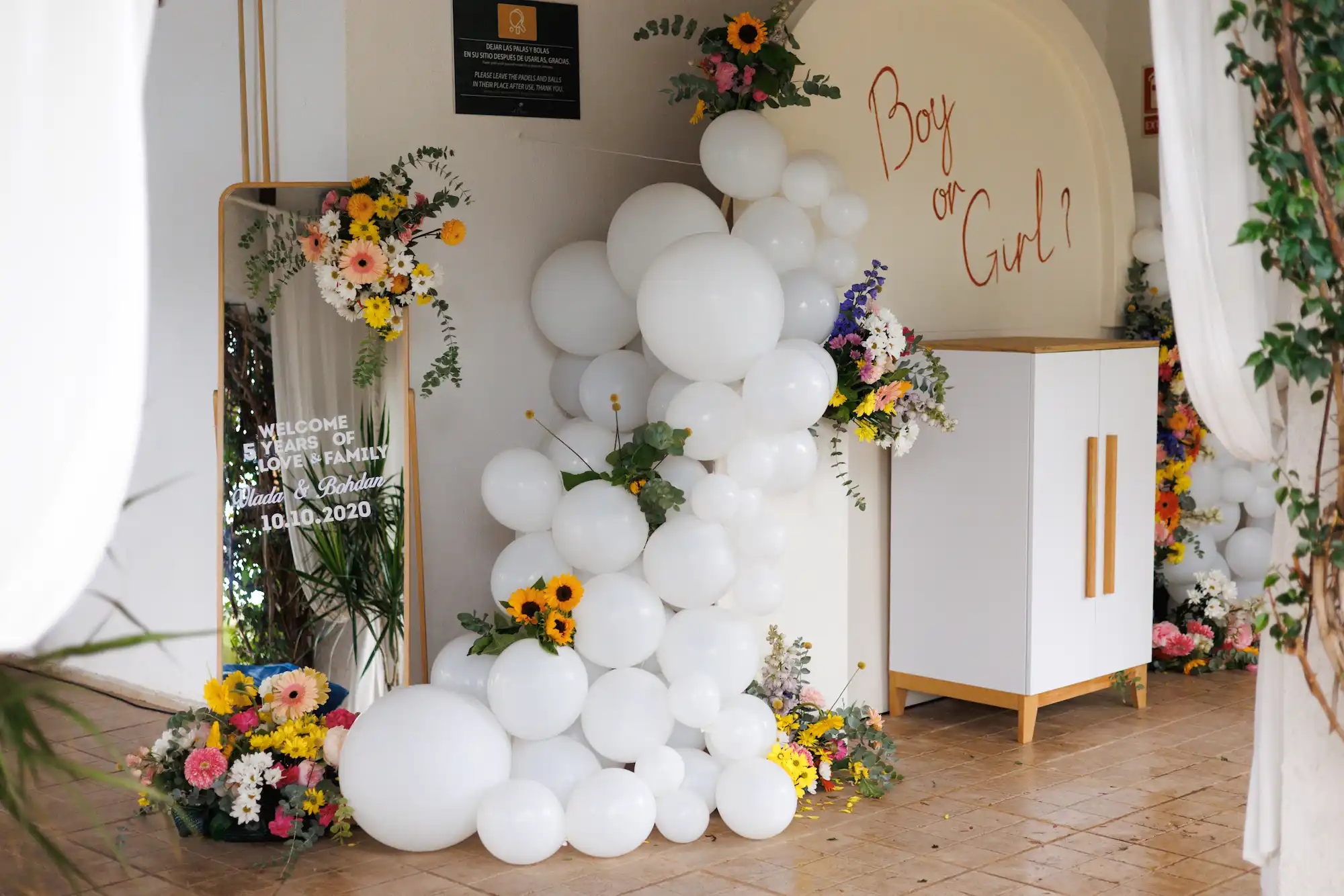 Decorative gender reveal setup with white balloons, colorful flowers, a mirror with 'Welcome 5 years of love & family Vlada & Bohdan 10.10.2020' text, and 'Boy or Girl?' written on a wall.