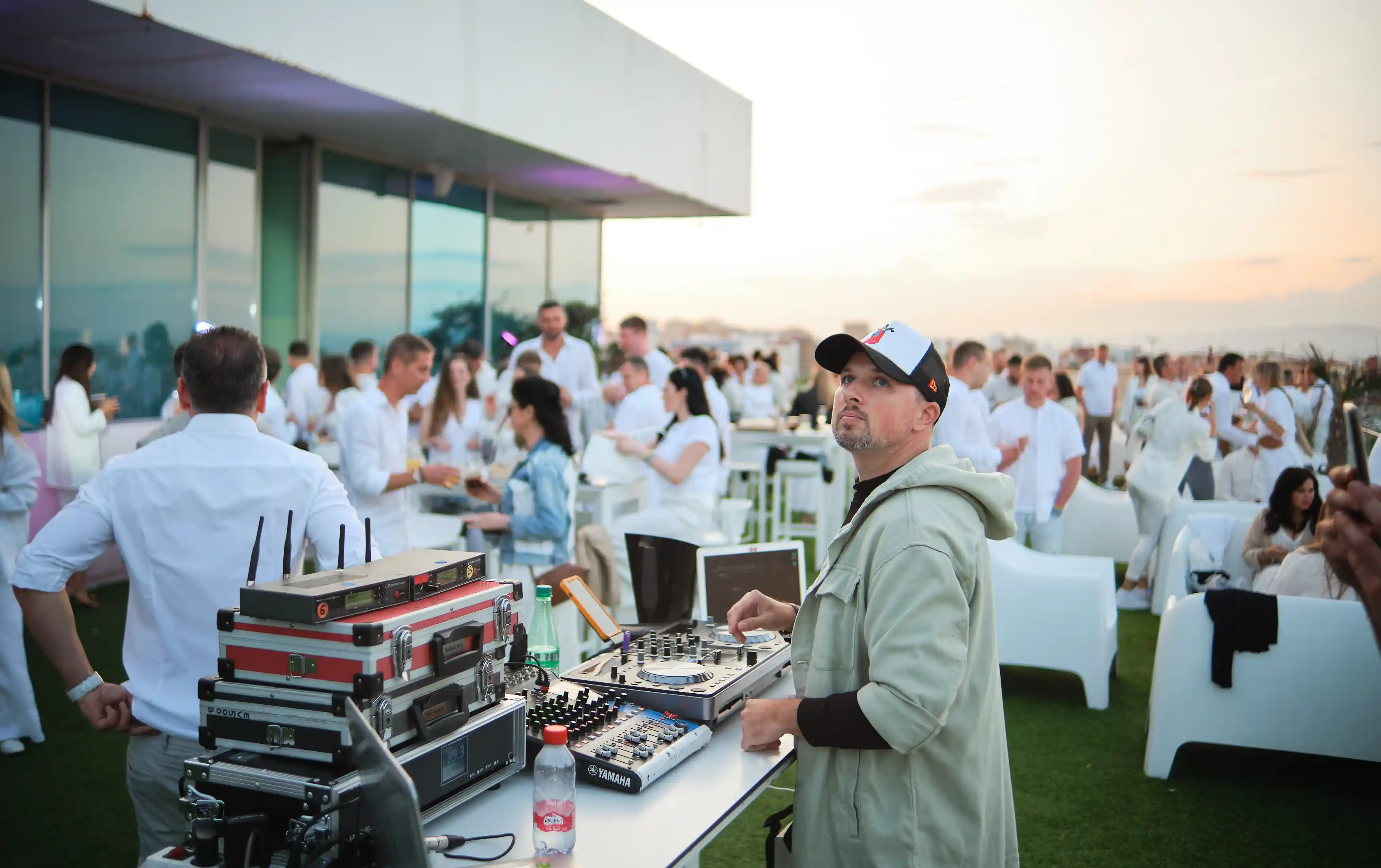 Ди-джей в кепке и светлой куртке стоит за оборудованием на вечеринке на открытом воздухе с гостями, одетыми преимущественно в белое.