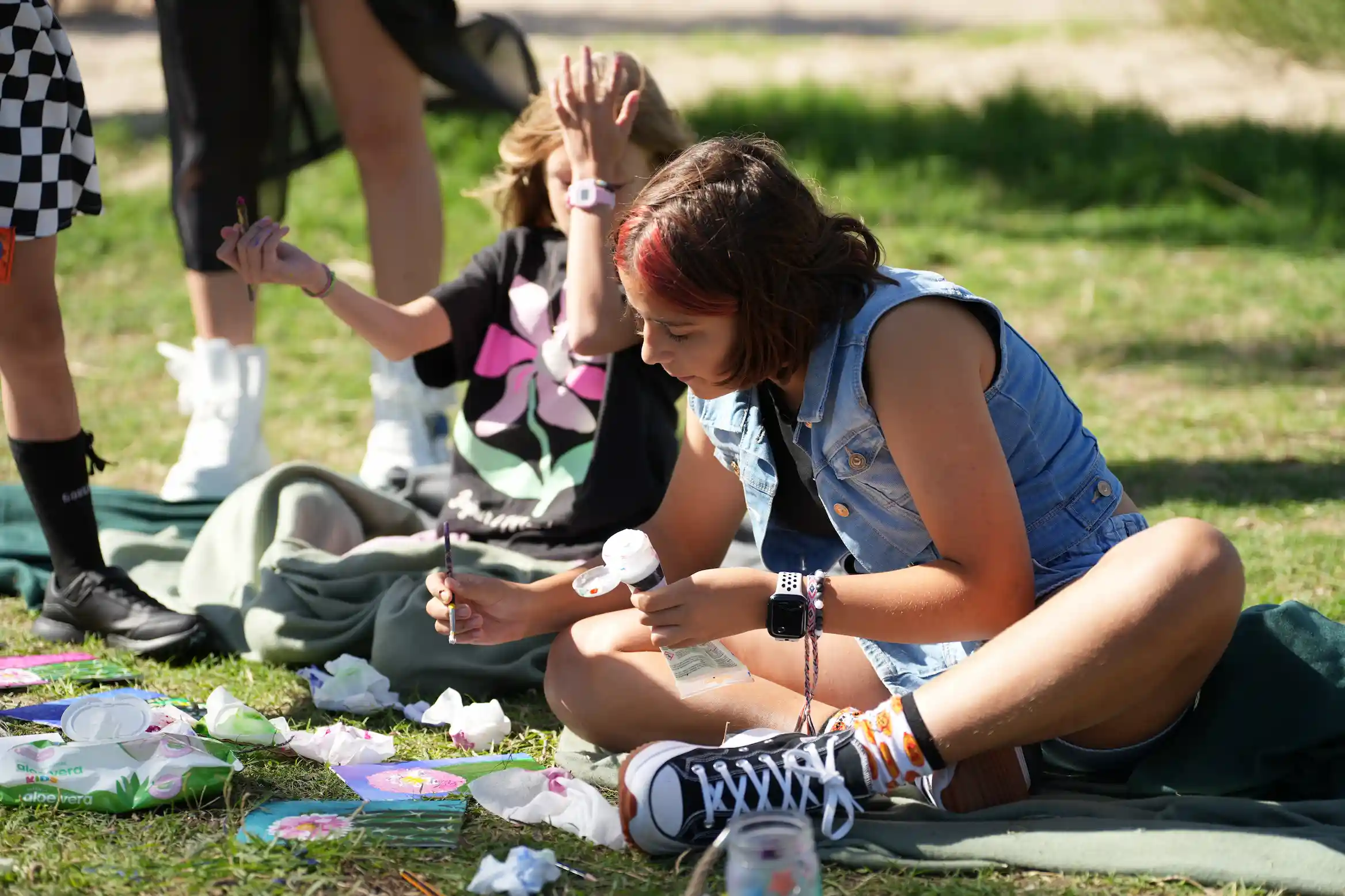 Two children sitting on grass outdoors painting on paper with art supplies around them.