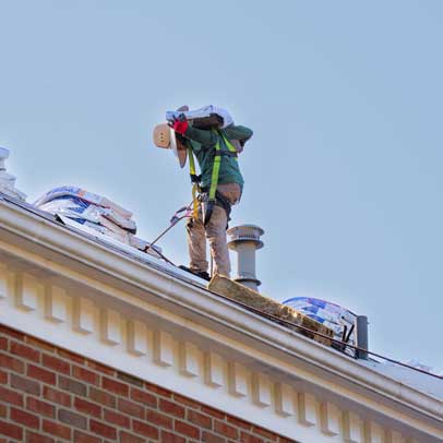 JF Baker roofer wearing safety harness, carrying a bag of roofing material on a house roof under clear sky.