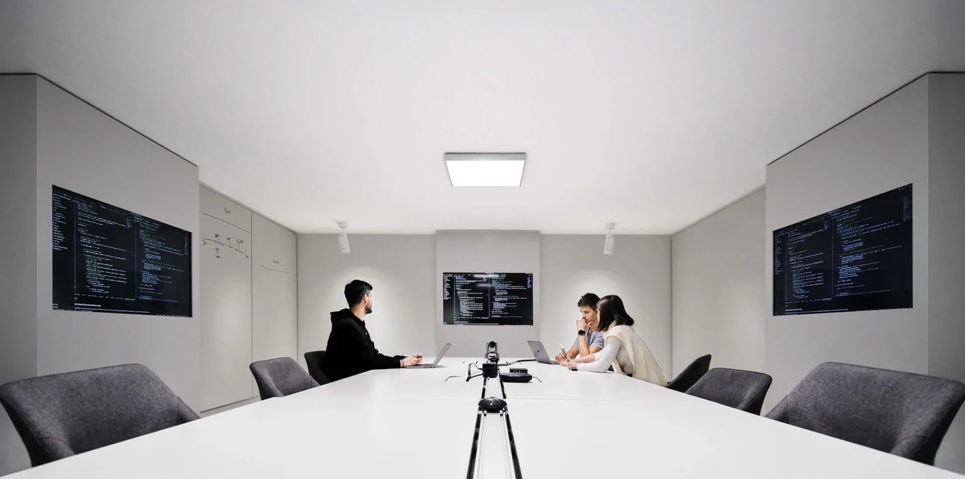 A modern conference room with multiple screens displaying code, where a group of developers is working collaboratively on laptops.