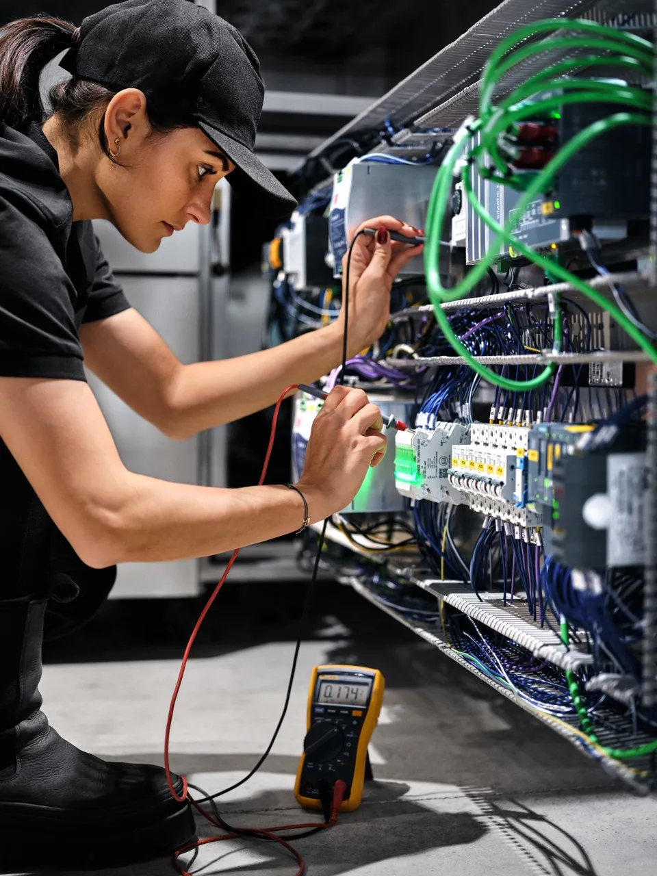 Technician testing electrical circuits inside an industrial control panel using a digital multimeter, surrounded by wires and components.