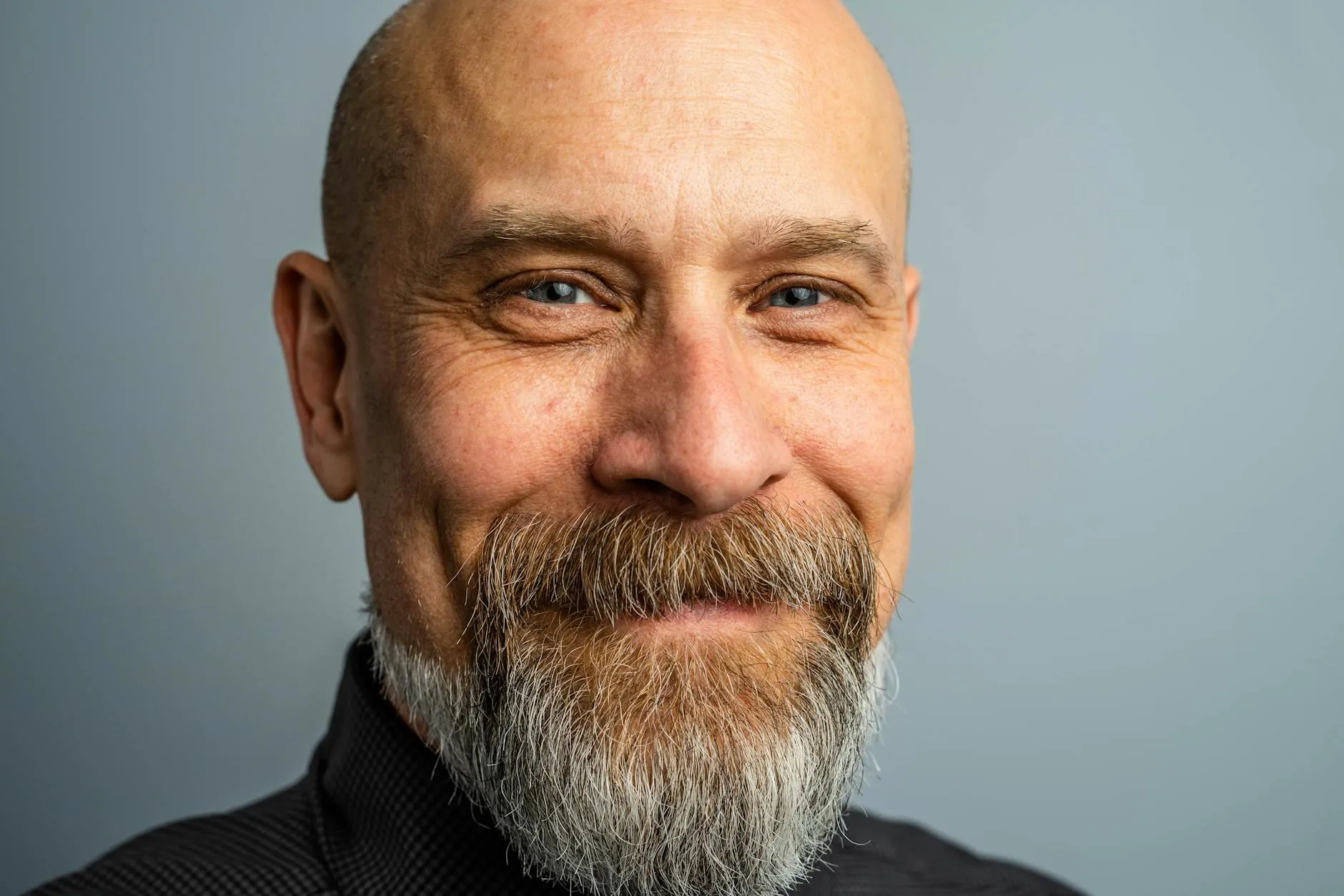Portrait of a smiling bald man with a grey beard, wearing a black shirt, against a plain light blue background.