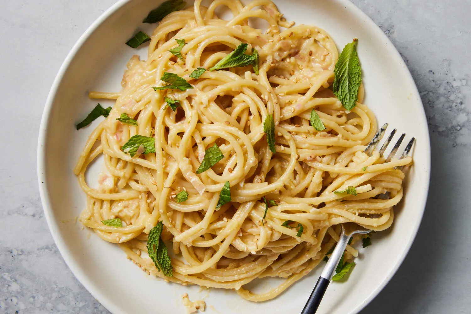 Top-down view of a plate of creamy spaghetti garnished with fresh herbs, with a fork resting on the side of the plate on a light stone surface.