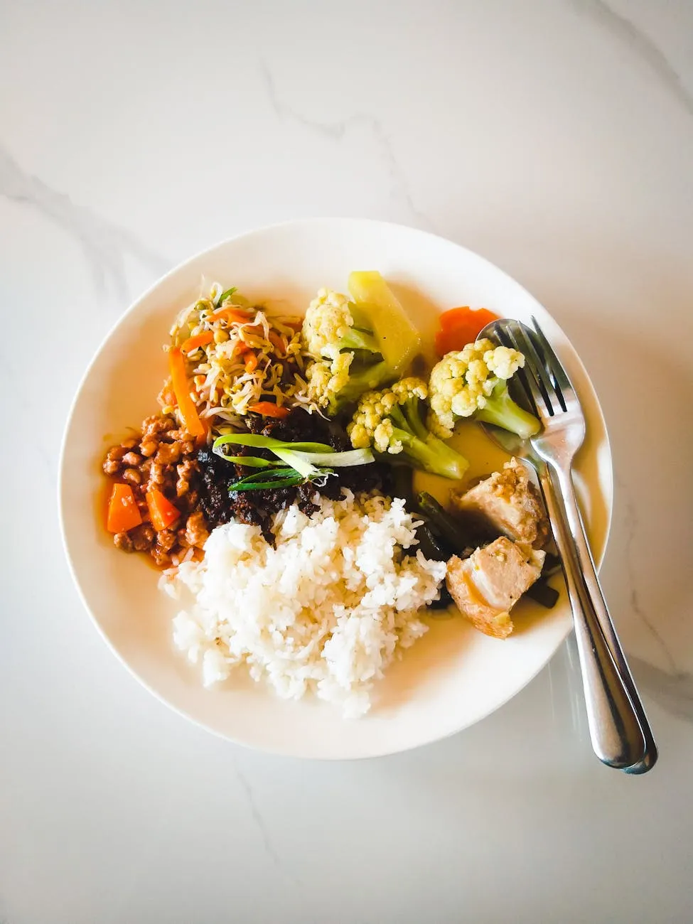 A colorful plate of food containing white rice, cooked vegetables, beans, and a portion of protein with a fork placed on the side, set on a clean white surface.