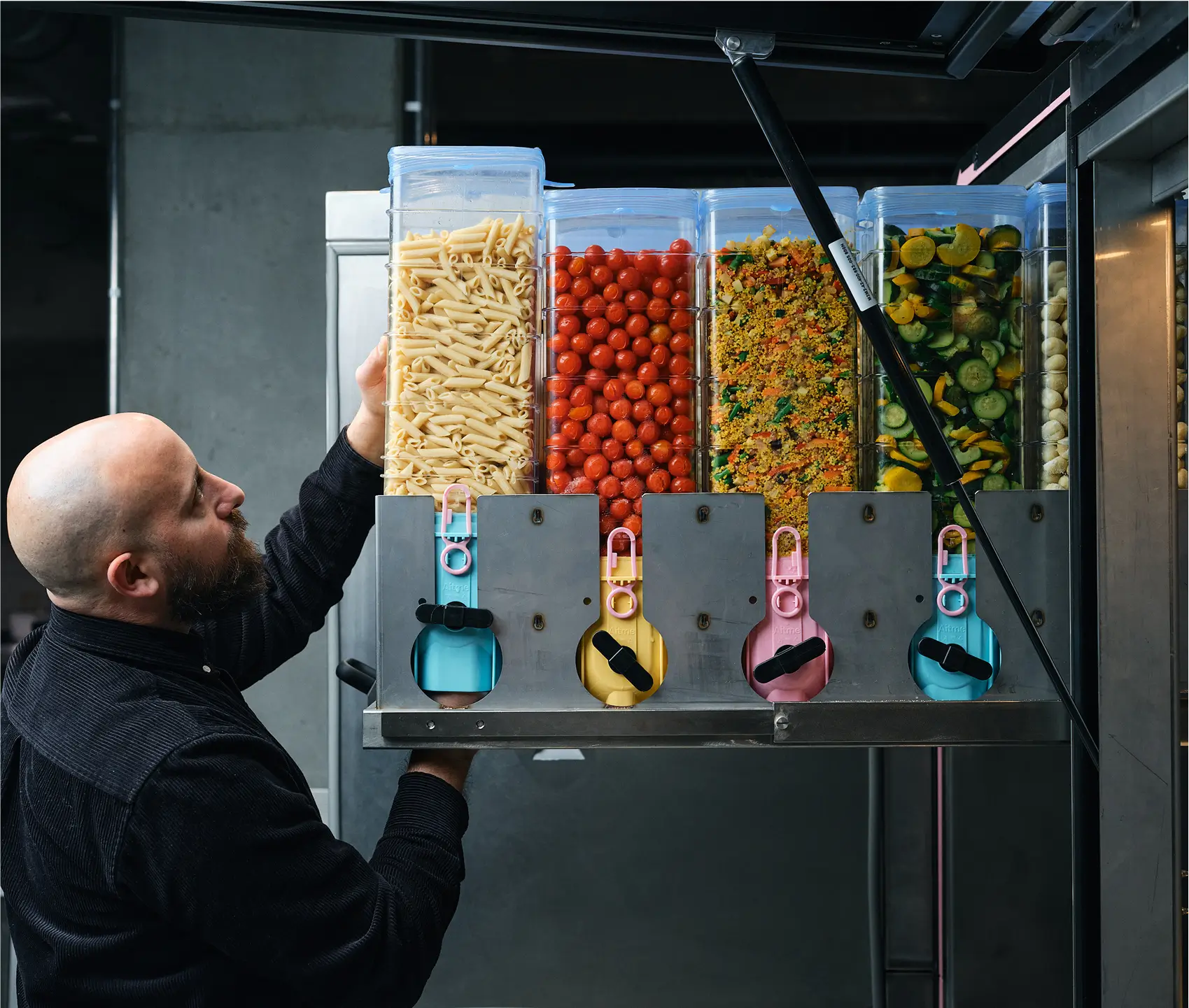 Operator refilling ingredient containers in a CIRCUS robotic kitchen system, featuring compartments filled with fresh food like pasta, cherry tomatoes, couscous, and sliced vegetables.