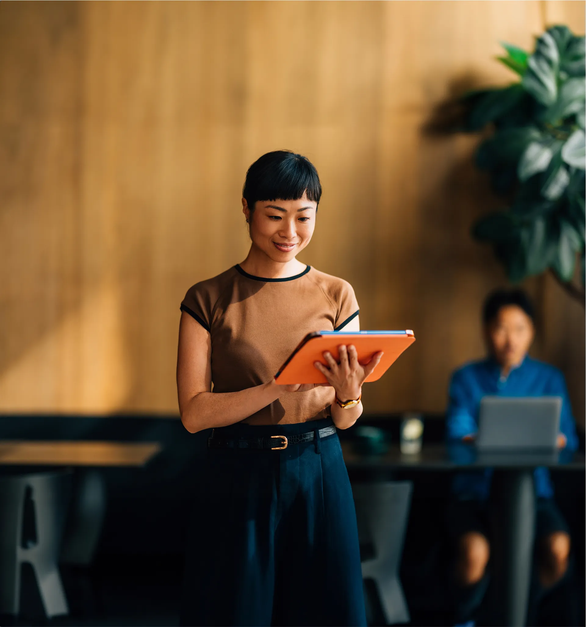 Smiling woman standing indoors holding an orange tablet, engaging with content, while another person works on a laptop in the background in a warmly lit modern workspace.