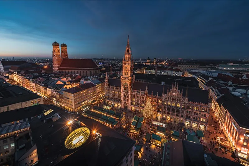 Aerial view of Munich’s Marienplatz at dusk during the Christmas season, with the New Town Hall, festive lights, market stalls, and the Frauenkirche in the background.