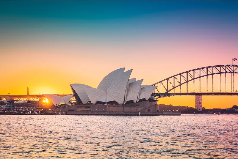 Sunset view of the Sydney Opera House with the Sydney Harbour Bridge in the background, glowing against a colorful evening sky.