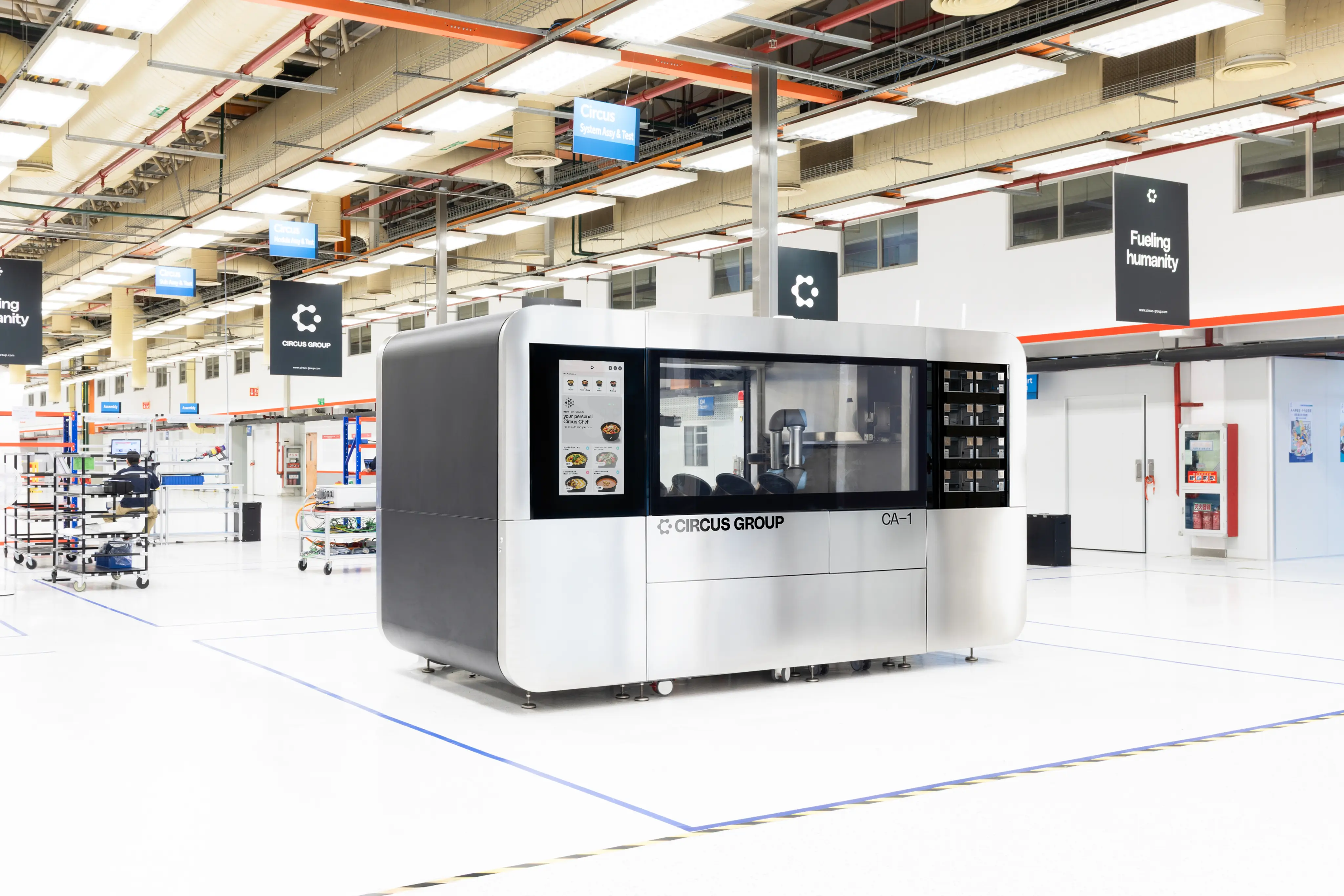 A large silver and black industrial machine labeled CIRCUS GROUP CA-1 inside a spacious, brightly lit factory with hanging signs and organized workstations.